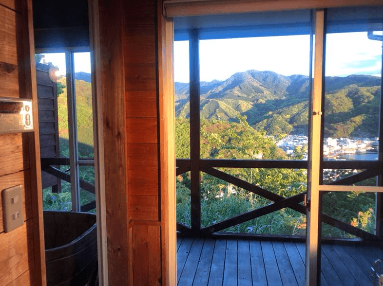 View of lush green mountains and a town from a wooden balcony with sliding glass doors.