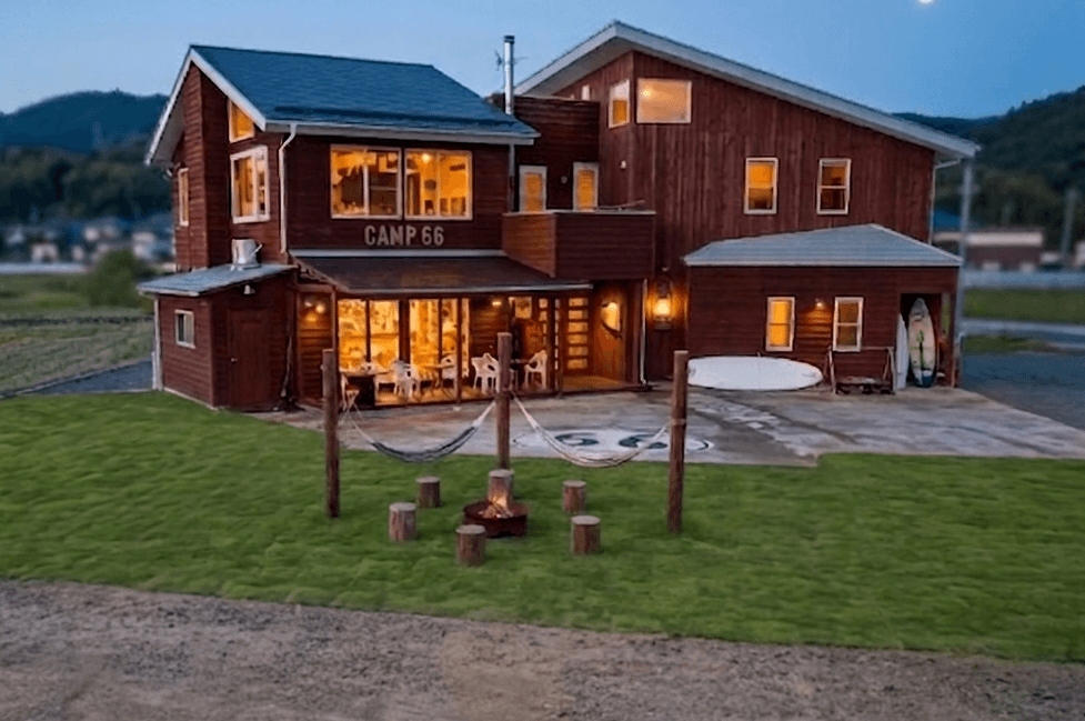 Two-story wooden lodge at dusk with a green lawn, fire pit, hammocks, and warm interior lighting.