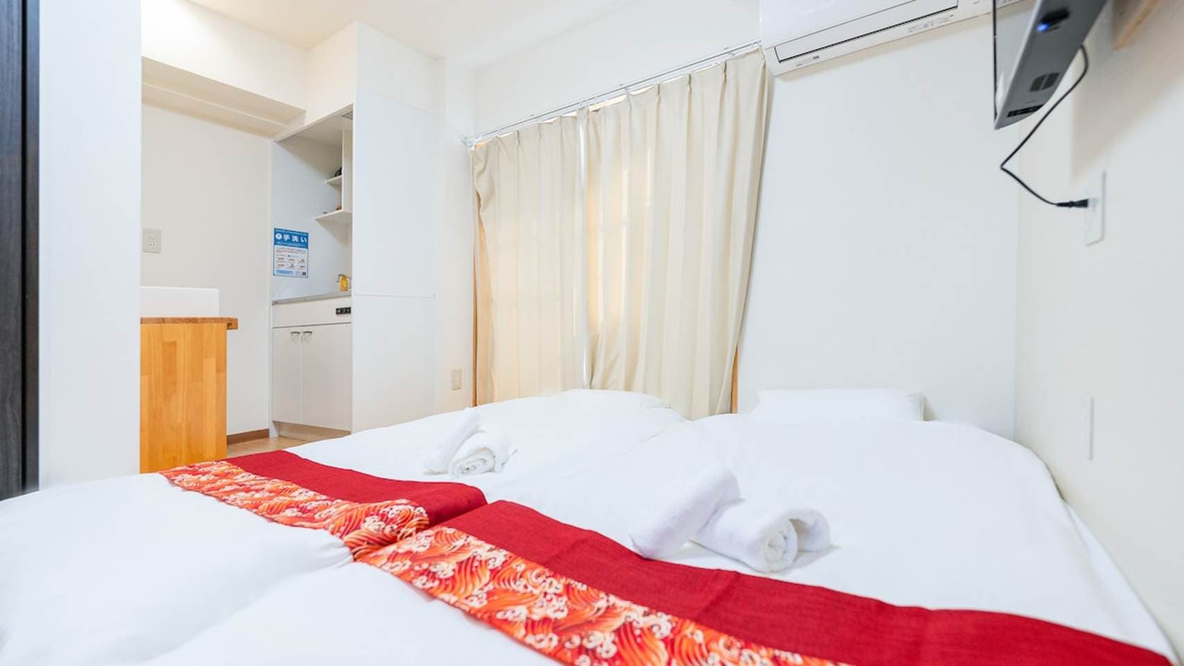 Beds with white linens and red patterned runners in a bright bedroom with beige curtains.
