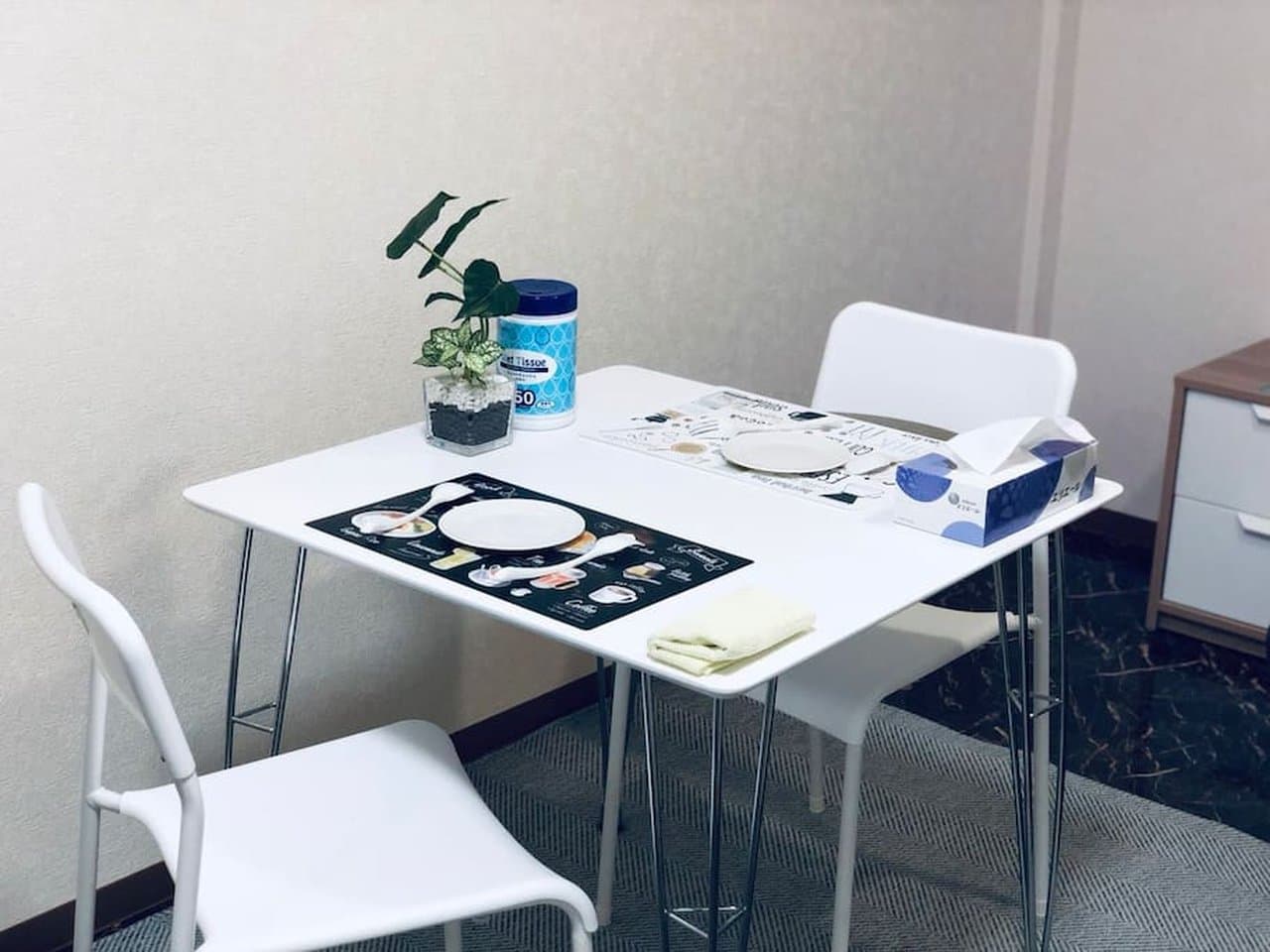 Small white dining table with two chairs, set with placemats, plates, and a small potted plant.