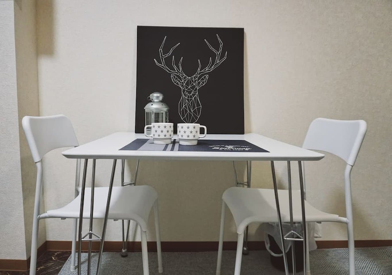 Small white dining table with two chairs, two mugs, and a geometric deer art print on the wall.
