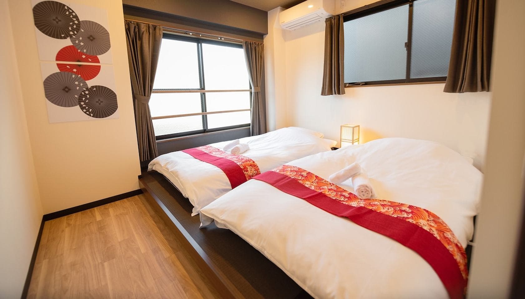 Two twin beds with red runners on a raised wooden platform in a bedroom with windows.