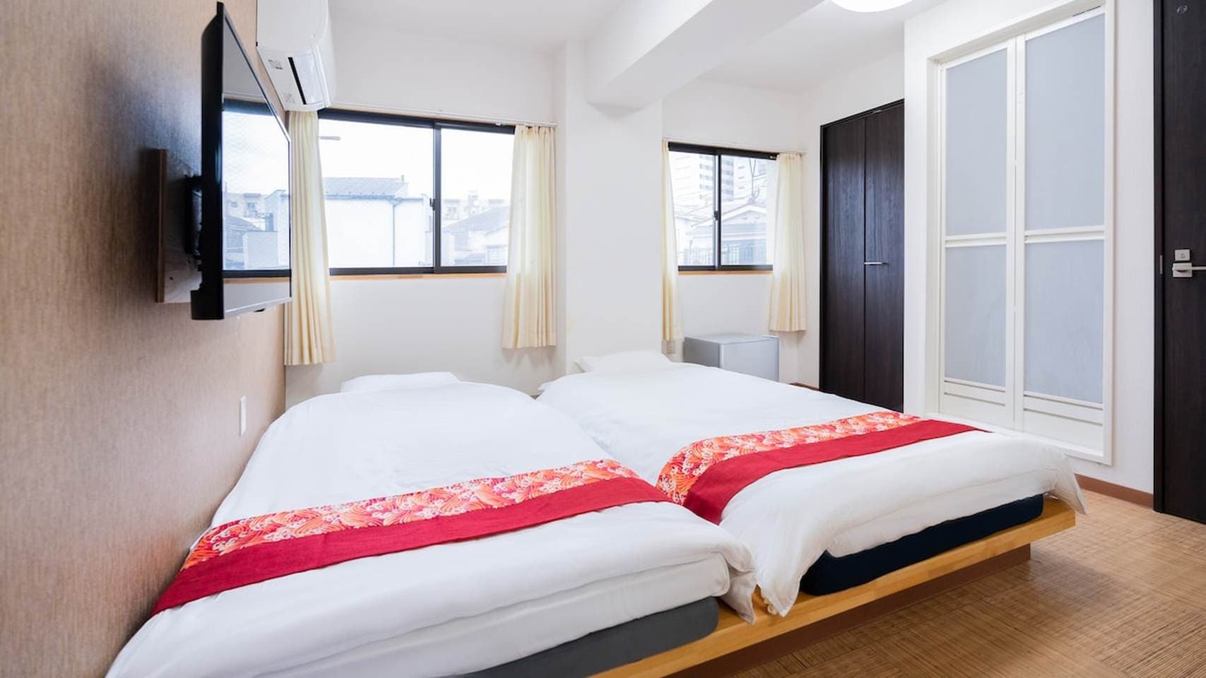 Bright bedroom with two twin beds, red bed runners, a wall-mounted TV, and windows.