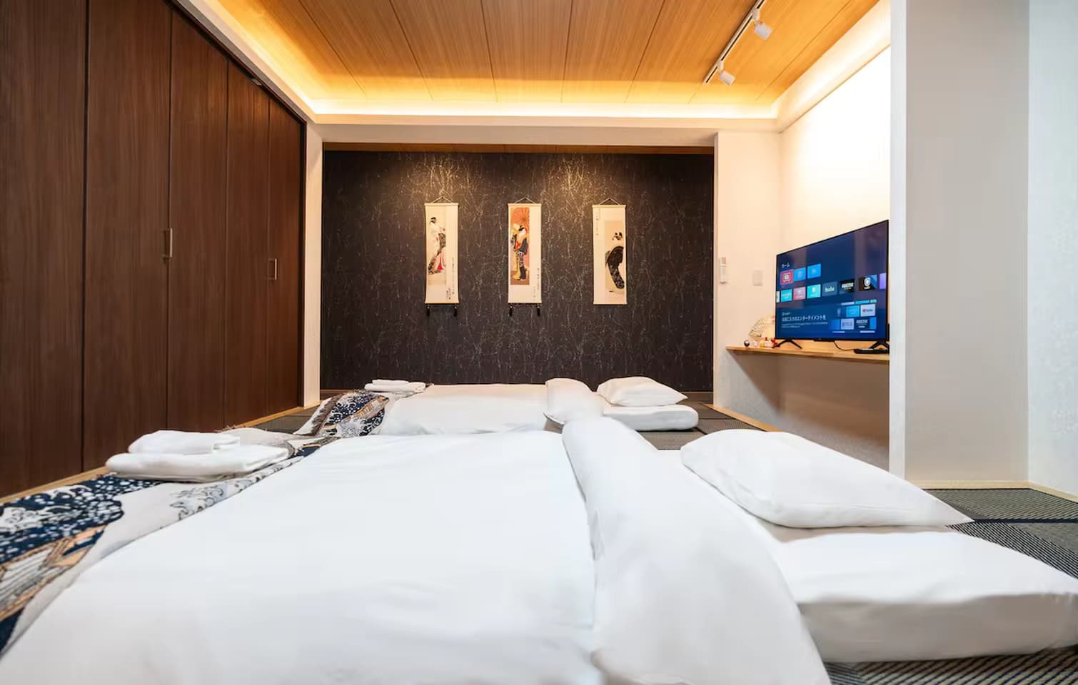 Japanese-style bedroom with futon bedding on tatami mats, hanging scrolls, and a television.