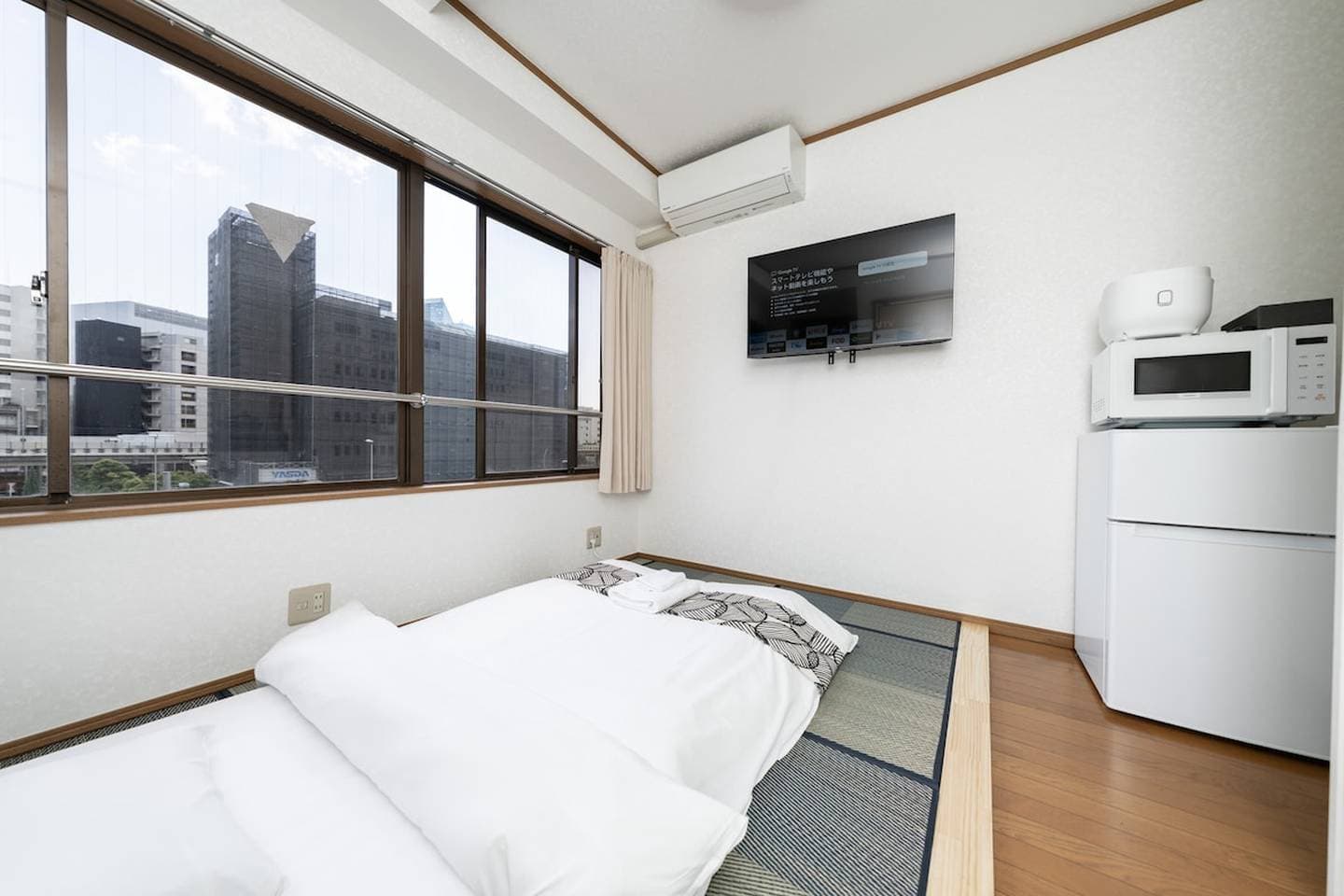 Japanese-style bedroom with a futon on tatami mats, a wall-mounted TV, and a window overlooking the city.
