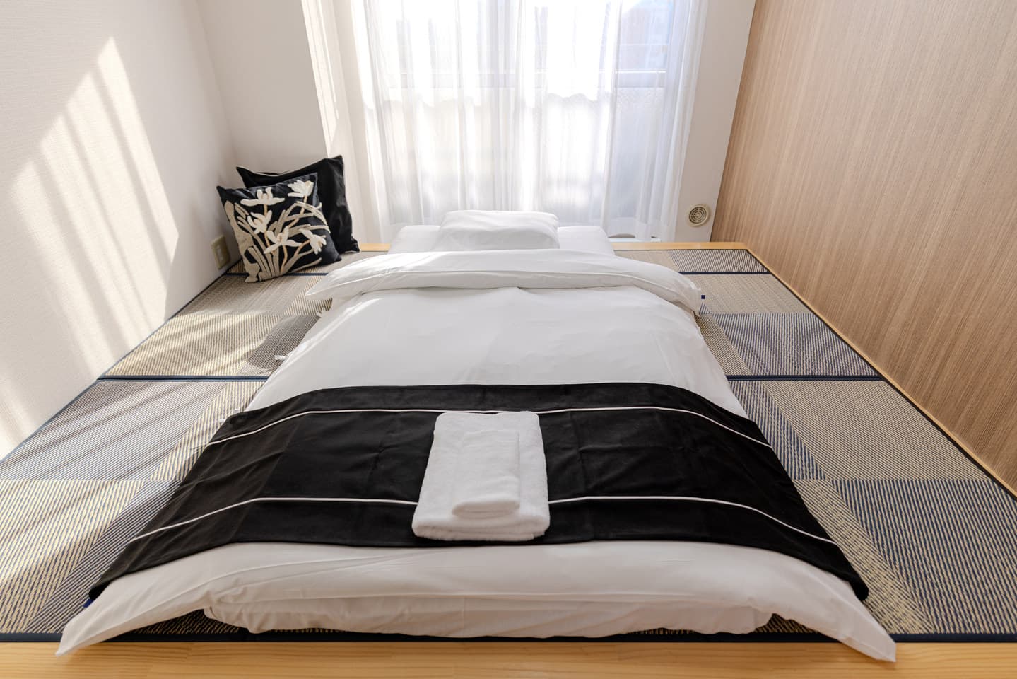 Japanese futon bed with white linens and a black runner laid out on tatami mats.