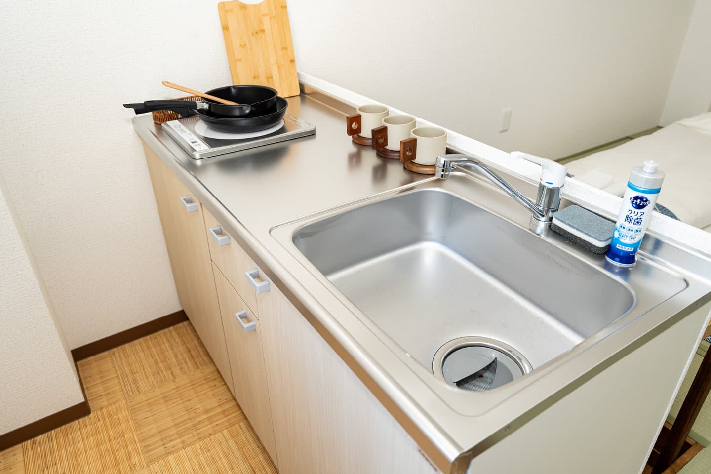 Compact kitchenette with a stainless steel sink, induction cooktop, frying pan, and mugs on a light wood counter.