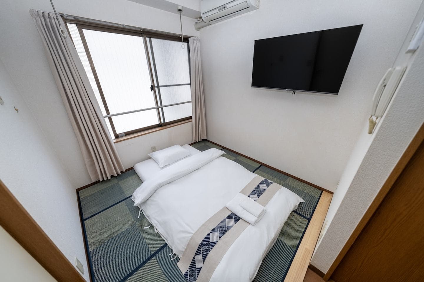 Japanese-style bedroom with a white futon on tatami mats, a wall-mounted TV, and a window with curtains.