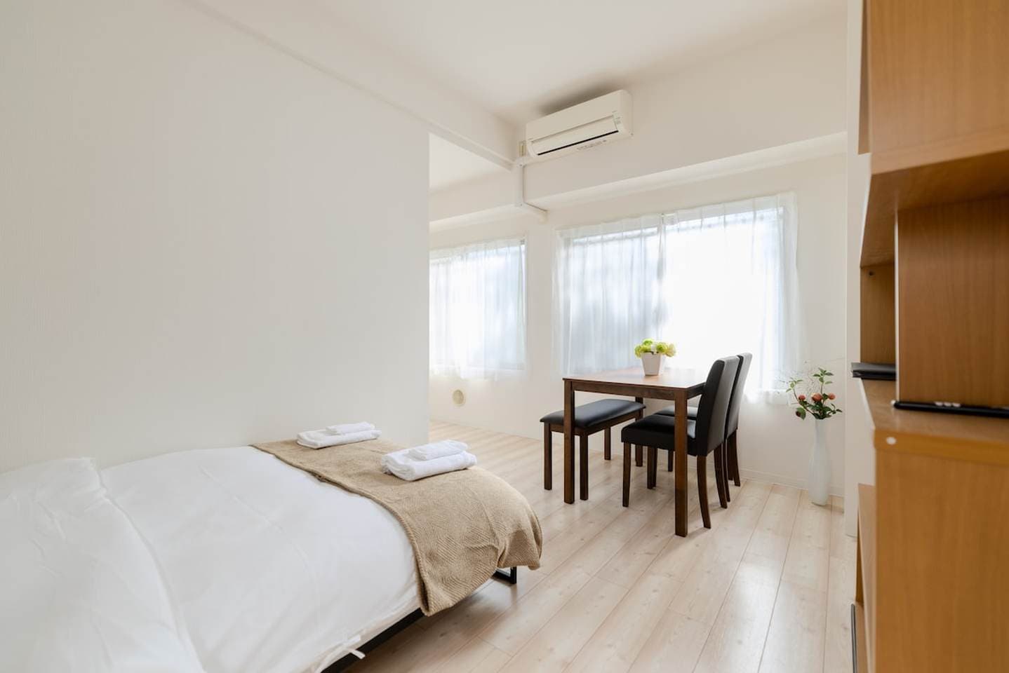 Bright studio apartment featuring a bed in the foreground and a wooden dining table set by the window.