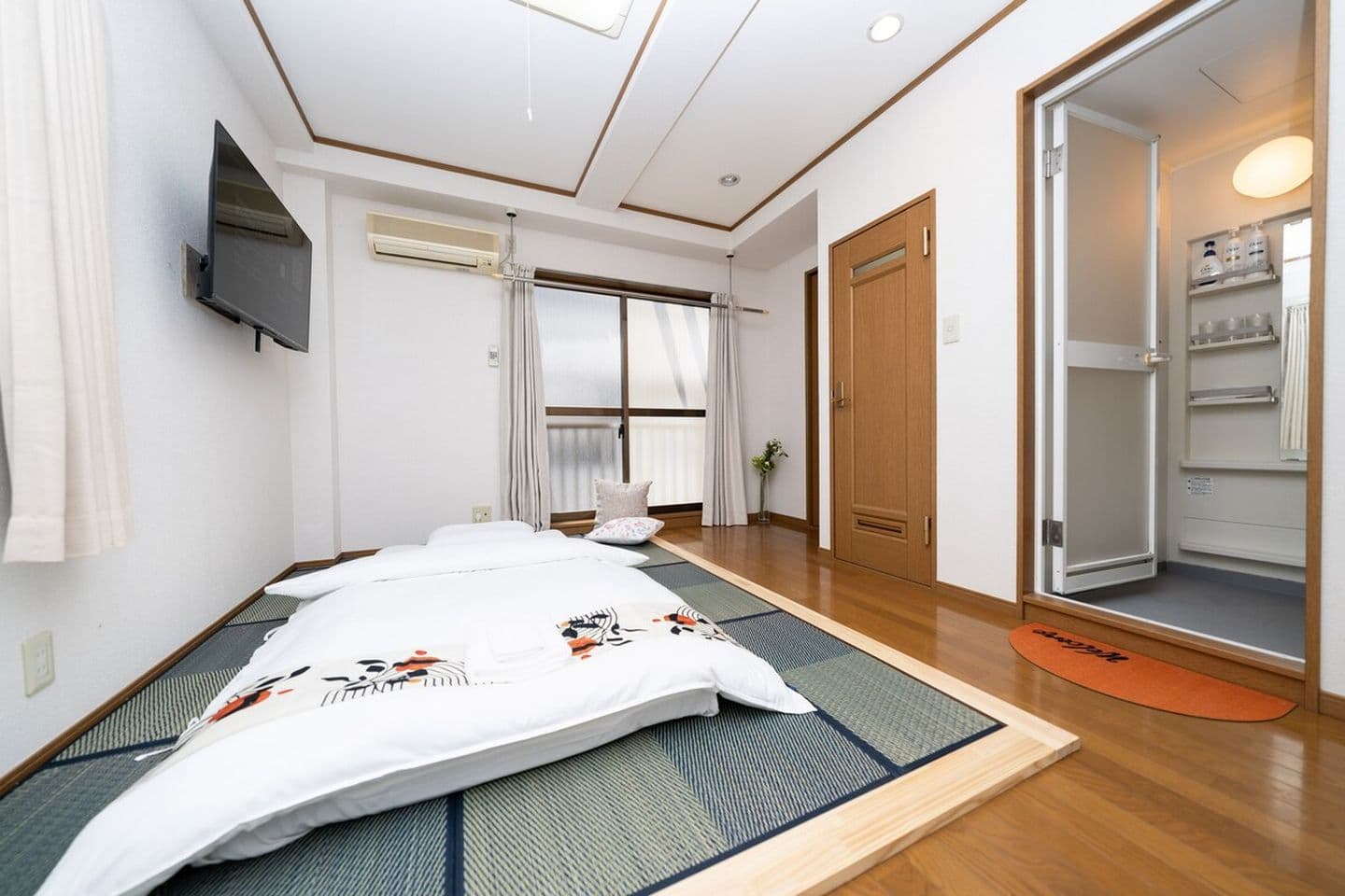 Japanese-style bedroom with a white futon on tatami mats, a wall-mounted TV, and an open door to the bathroom.