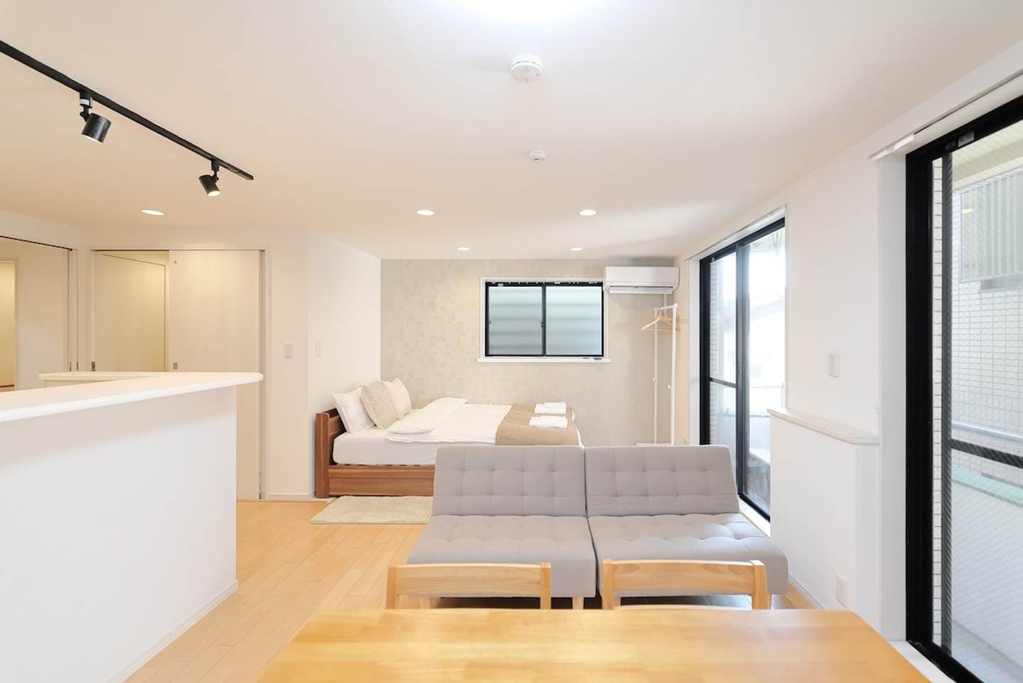 Bright studio apartment with a grey sofa, dining table, and a double bed in the background.