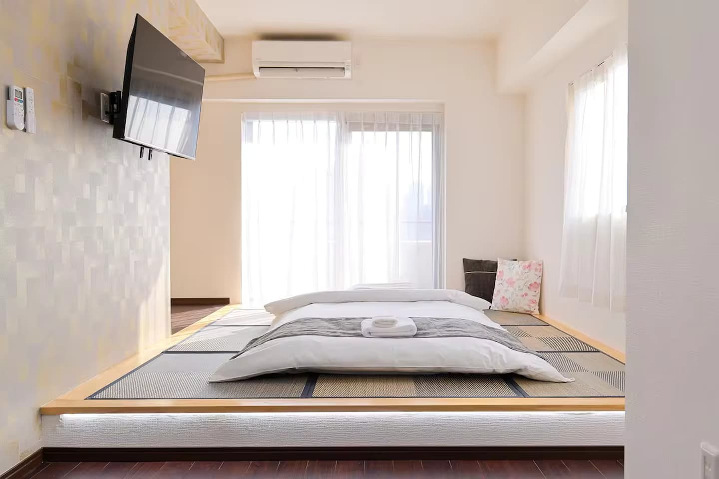 Futon bed on a raised tatami platform in a bright room with a wall-mounted TV and sheer curtains.