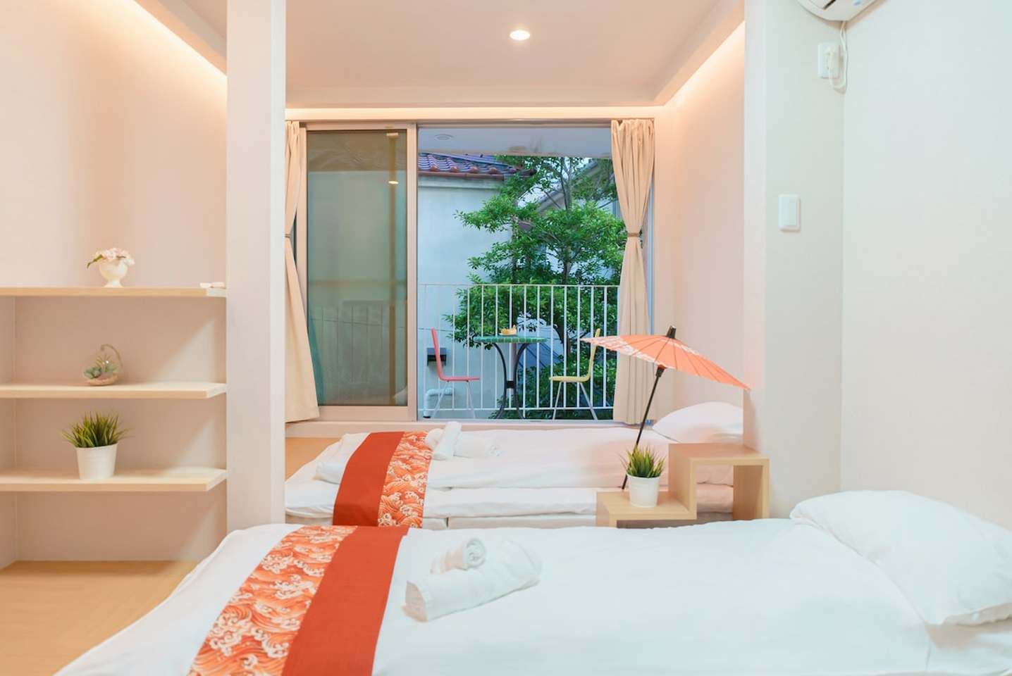 Minimalist bedroom with two single beds, wooden shelving, and a sliding door opening to a balcony.
