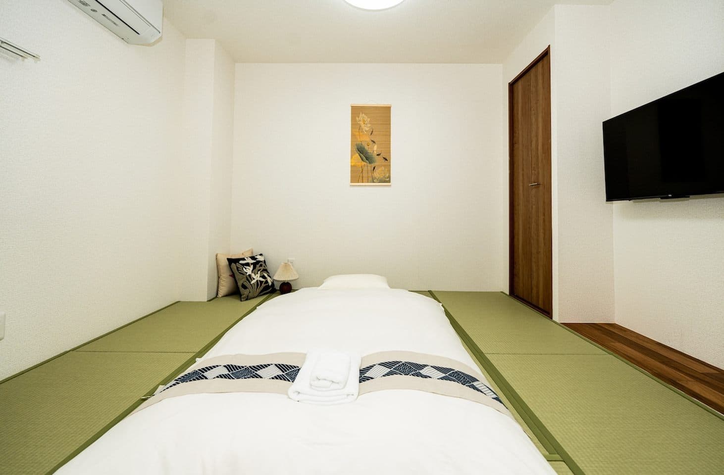 Futon bed laid out on green tatami mats in a minimalist Japanese-style bedroom with a wall-mounted TV.