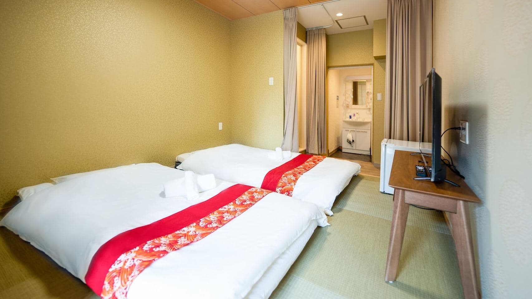 Japanese-style bedroom with two futon beds on tatami mats, a TV on a wooden stand, and a sink in the background.