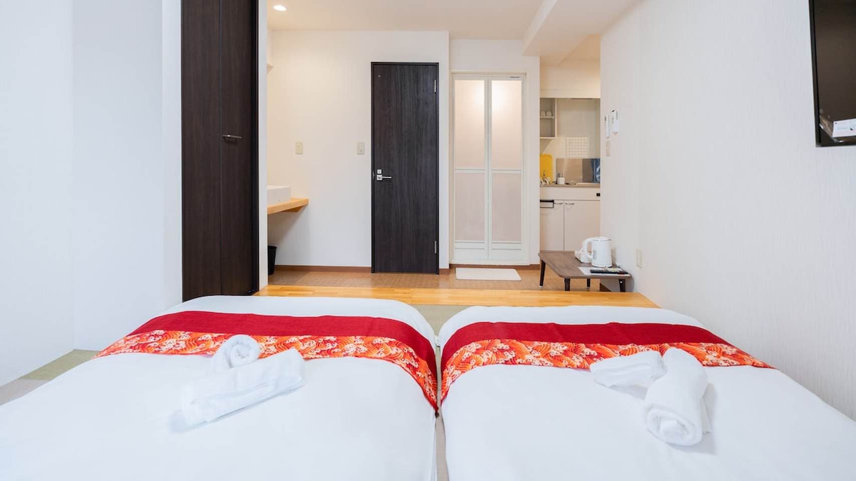Two low beds with red patterned runners and rolled towels in a modern studio apartment.