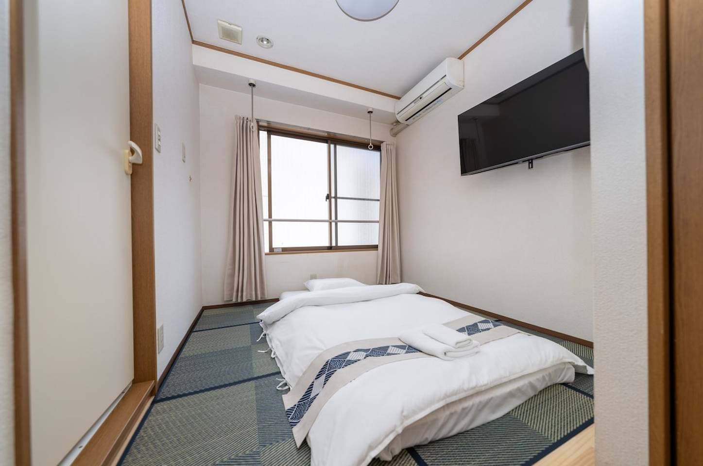 Japanese-style bedroom with a futon on tatami flooring, a wall-mounted TV, and a window.