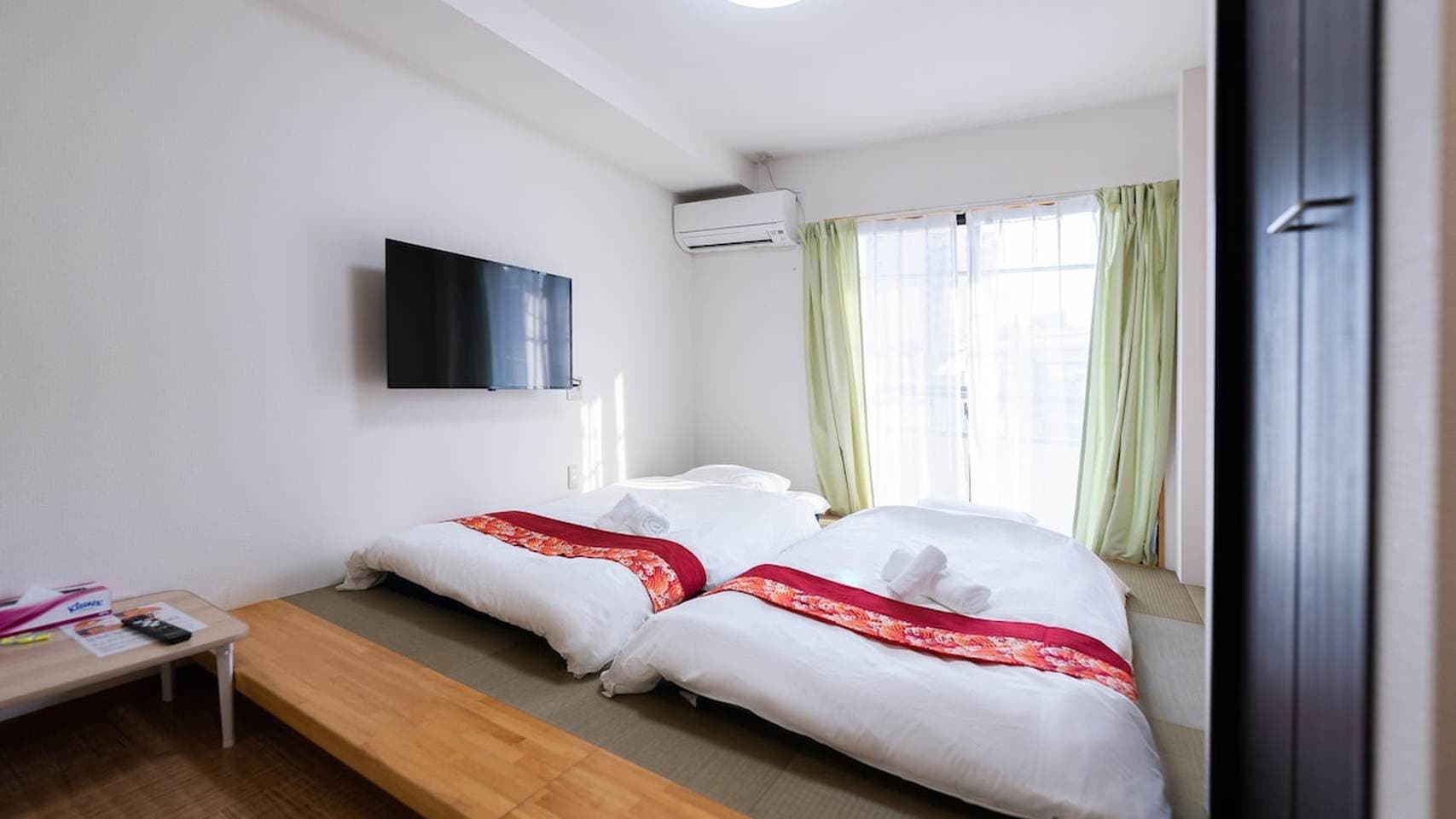 Two futon beds on a tatami platform in a bedroom with a wall-mounted TV and light green curtains.