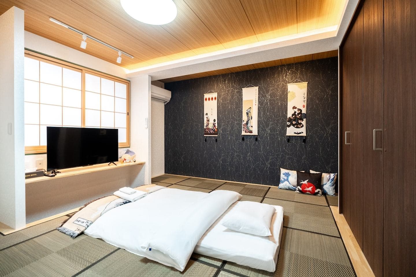 Japanese-style bedroom with futon bedding on tatami mats, a flat-screen TV, and decorative hanging scrolls.