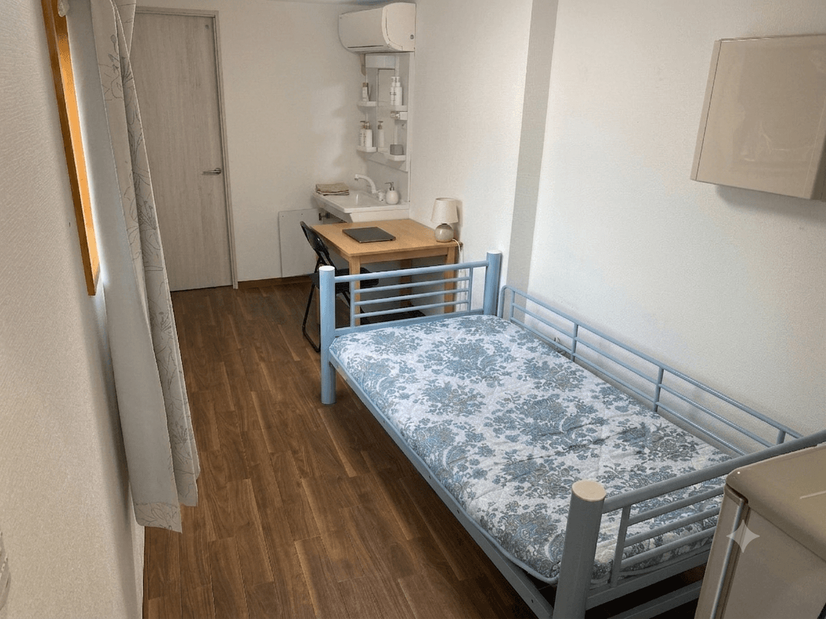 Compact bedroom featuring a blue metal single bed, a wooden desk, and a vanity sink.