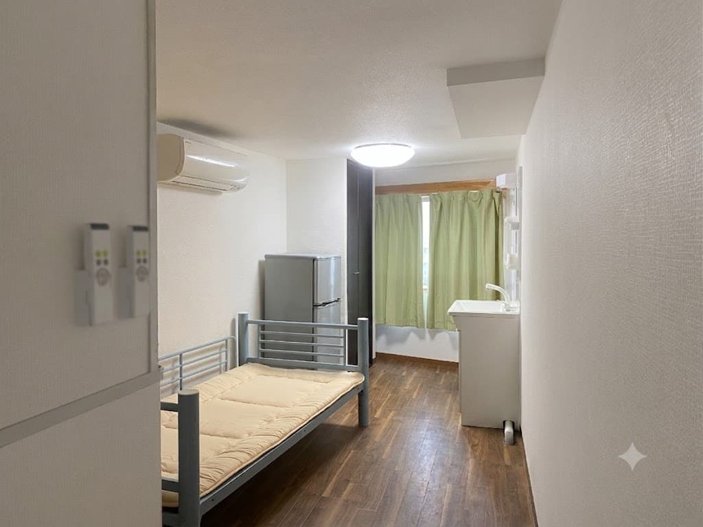 Compact studio room featuring a single bed, refrigerator, sink unit, and window with green curtains.