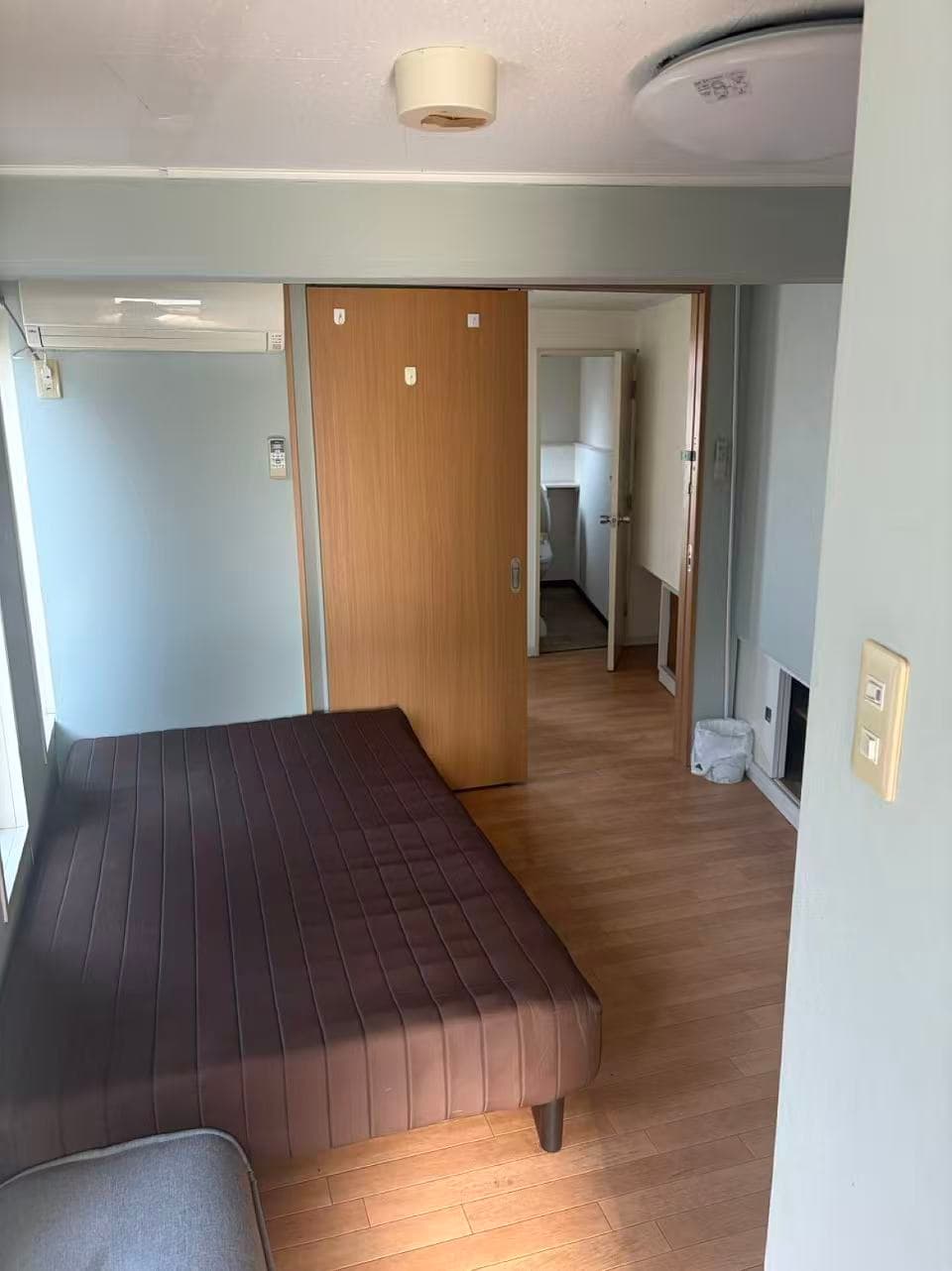 Compact bedroom with a brown mattress, wooden flooring, and a sliding door leading to a hallway.