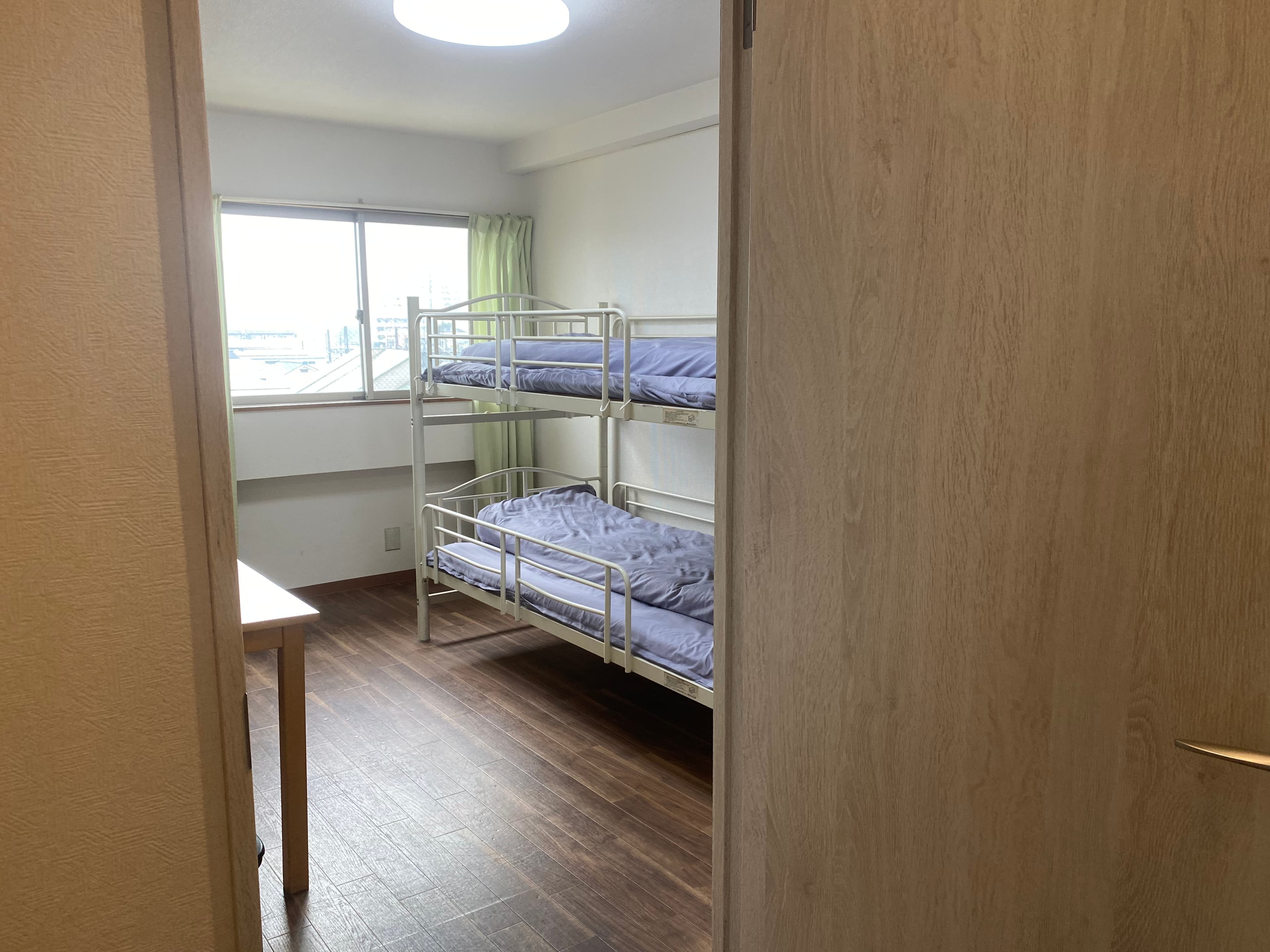 Bedroom with a white metal bunk bed, wooden flooring, and a window with green curtains.