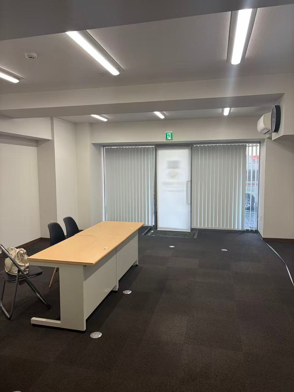 Office workspace with a wooden desk, chairs, dark carpet, and vertical blinds on glass doors.