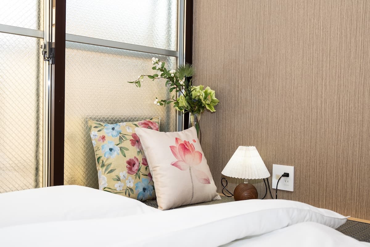 Bedside setup with floral pillows, a small pleated lamp, and a vase of flowers next to a window.