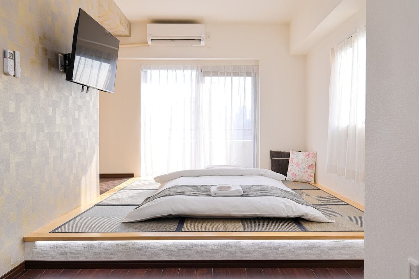 Futon bed on a raised tatami platform in a bright room with a wall-mounted TV and sheer curtains.