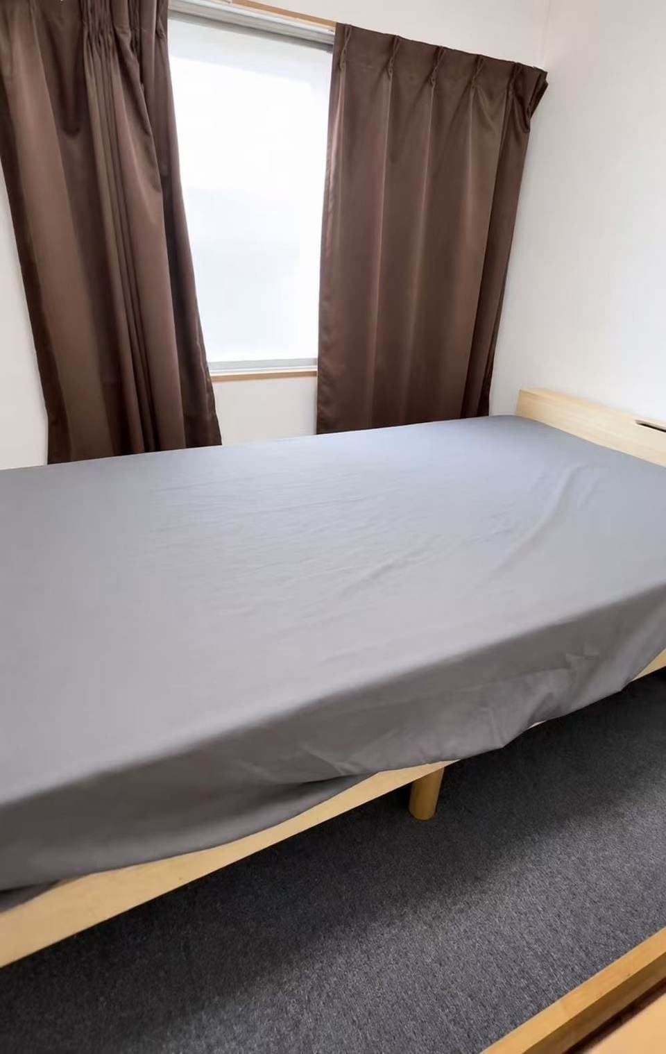 A bed with a grey sheet and wooden frame in front of a window with brown curtains.