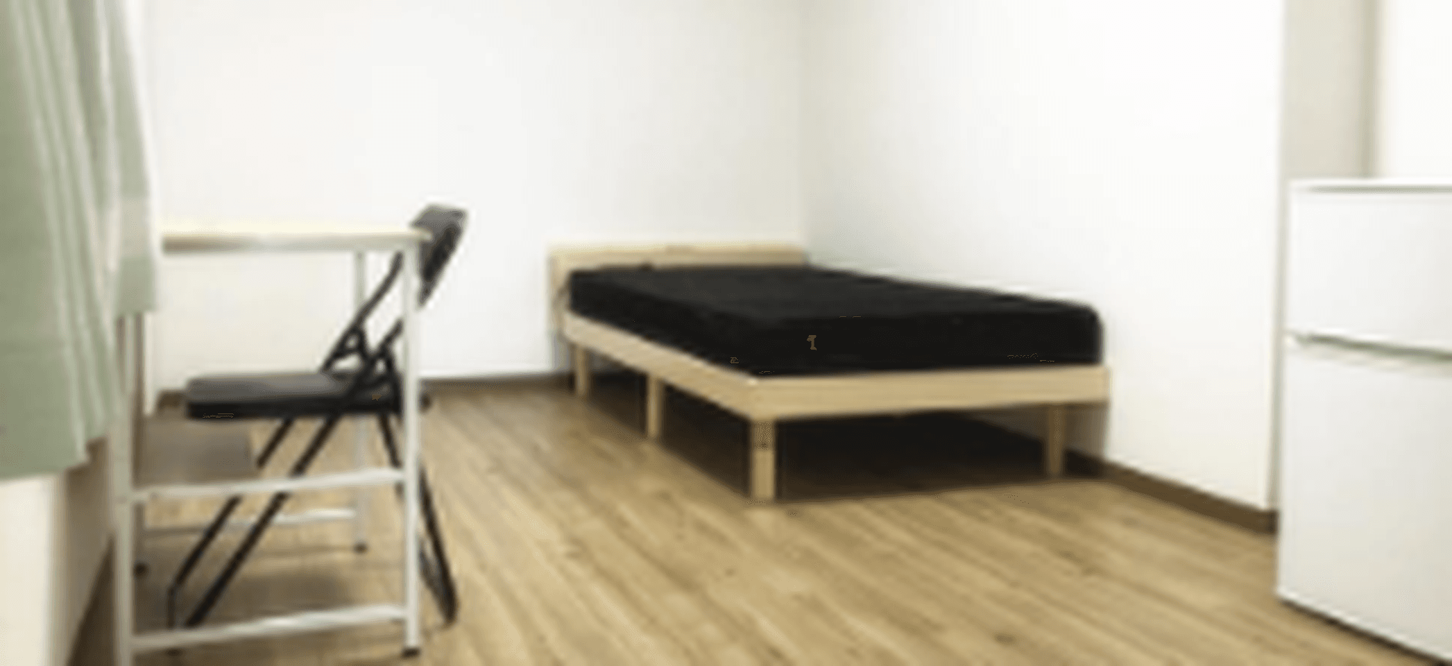 Minimalist bedroom with a single bed, a small desk and chair, and a mini fridge on wood flooring.