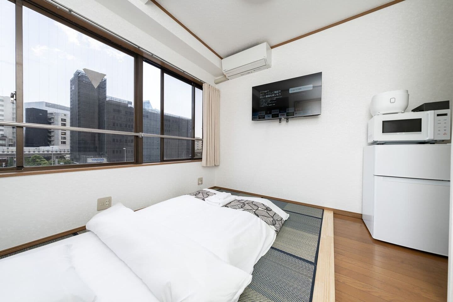 Japanese-style bedroom with a futon on tatami mats, a wall-mounted TV, a mini fridge, and a large window.