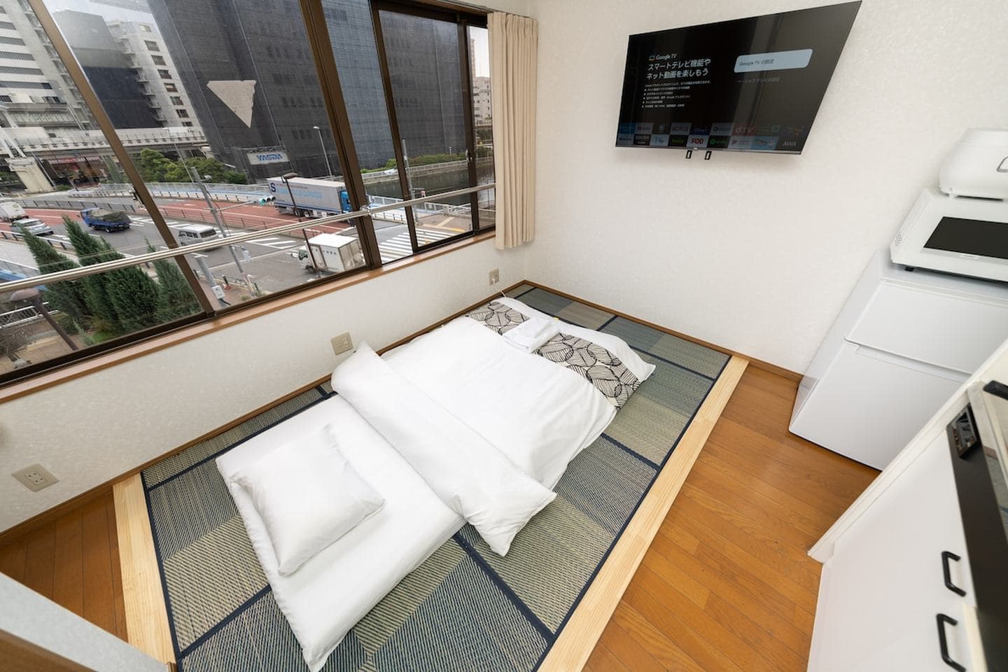 Japanese futon bed on tatami mats in a studio apartment with a wall-mounted TV and city view window.