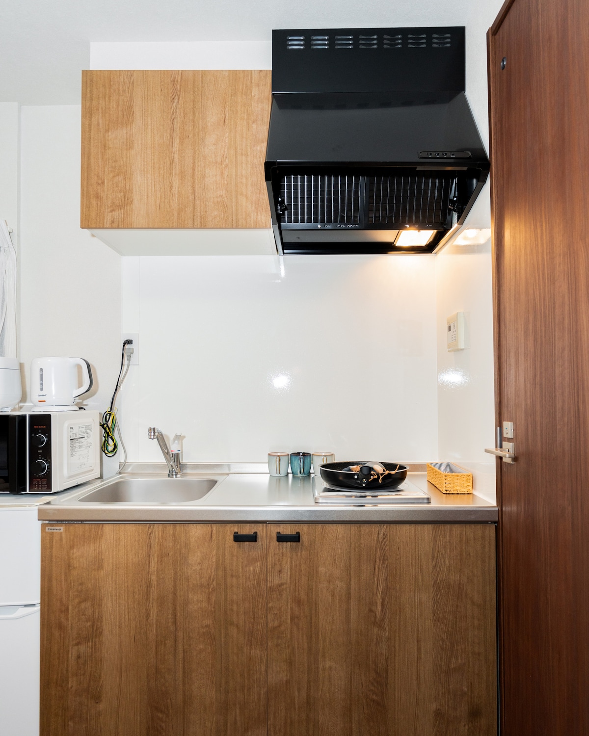 Compact kitchenette with wooden cabinets, stainless steel sink, electric stove, and black range hood.