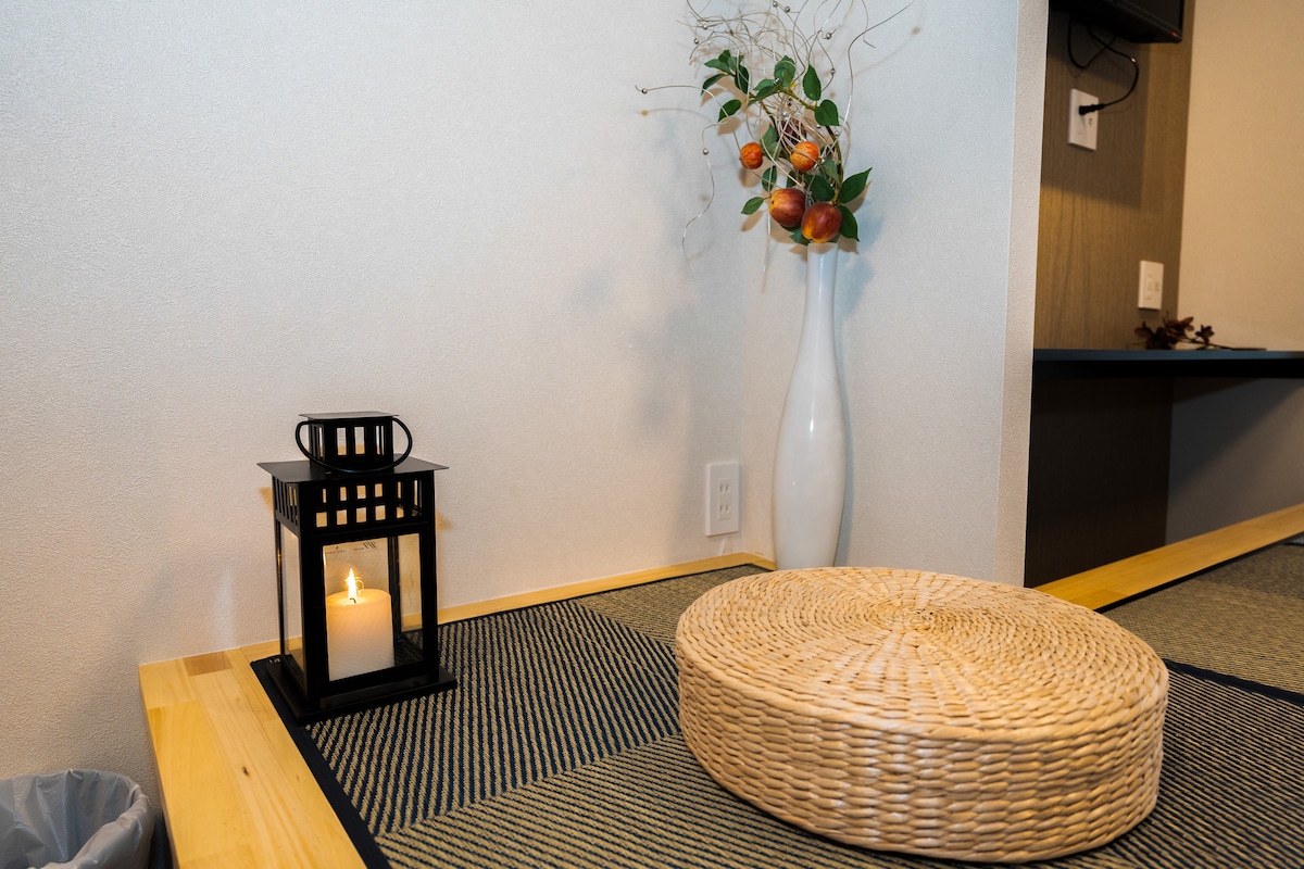 Woven straw cushion, black candle lantern, and a white vase with branches on tatami mat flooring.